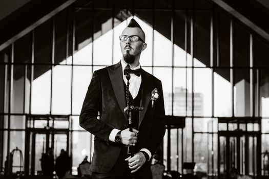 The groom in a tuxedo and bow tie with a mohawk hairstyle and a cane in the interior . black and white photo.