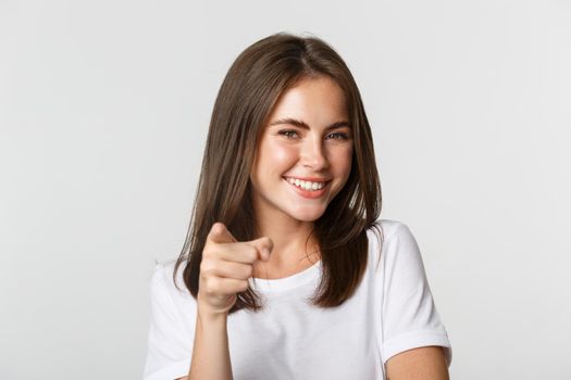 Close-up of happy smiling brunette girl pointing at camera, standing white background.