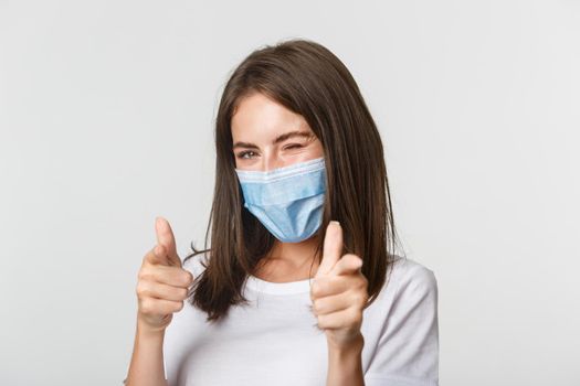 Covid-19, health and social distancing concept. Sassy smiling brunette girl in medical mask winking flirty at camera and pointing fingers.