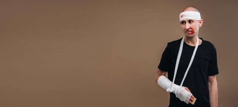 A battered man with a bandaged head and a cast on his arm stands on crutches on a gray background.