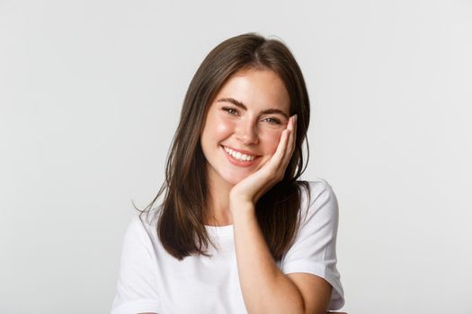 Close-up of beautiful smiling brunette girl touching cheeks, blushing over white background.