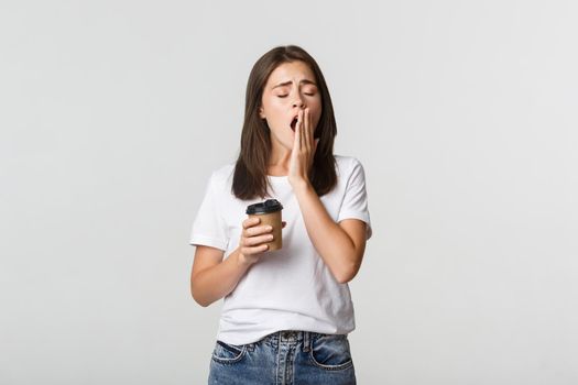 Portrait of tired and sleepy young attractive girl yawning while drinking takeaway coffee, white background.