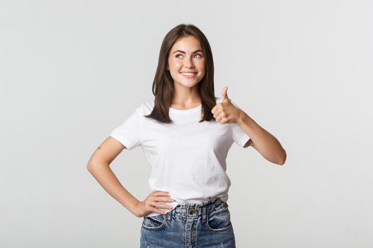 Satisfied smiling young woman showing thumbs-up in approval, looking upper left corner.