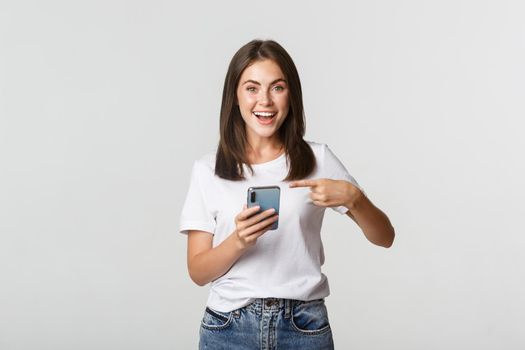Excited and pleased brunette girl pointing finger at smartphone screen.