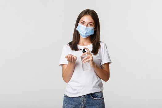 Covid-19, health and social distancing concept. Attractive young brunette woman in medical mask applying hand sanitizer on hand, white background.