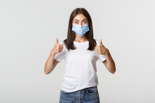 Covid-19, health and social distancing concept. Impressed young girl in medical mask showing thumbs-up in approval.