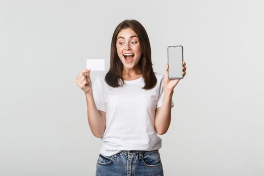 Excited attractive girl showing smartphone screen and credit card, white background.
