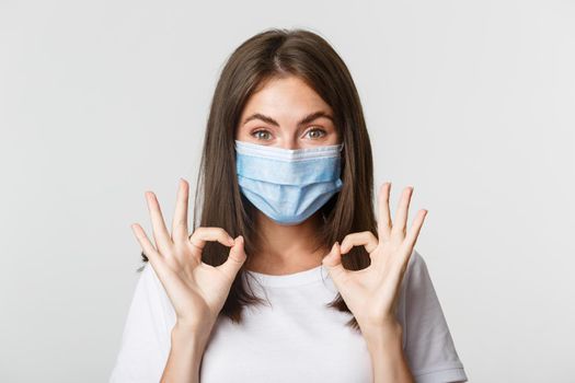 Covid-19, health and social distancing concept. Close-up of pretty satisfied girl in medical mask showing okay gesture in approval.