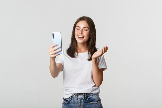 Friendly attractive girl video calling friend, smiling and having conversation, holding smartphone, white background.
