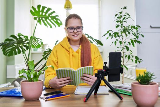 Teenage girl looking at webcam using smartphone for an online lesson, reading textbook book. Student 12, 13 years old, studying remotely at home, e-learning, virtual lessons, distance learning