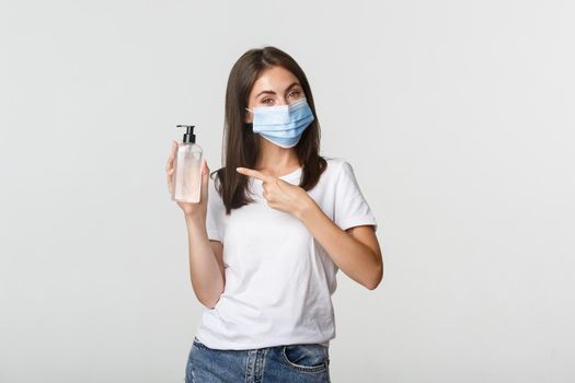 Covid-19, health and social distancing concept. Smiling cute brunette woman in medical mask advice using hand sanitizer, pointing at bottle.
