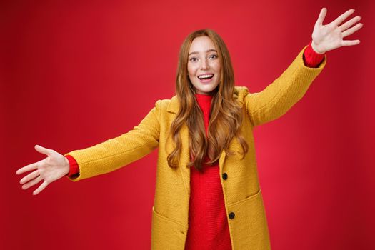 Hey come and hug me friend. Charming playful and emotional redhead female extanding palms forward to cuddle and greet, giving warm welcome smiling broadly posing in yellow coat over red wall.