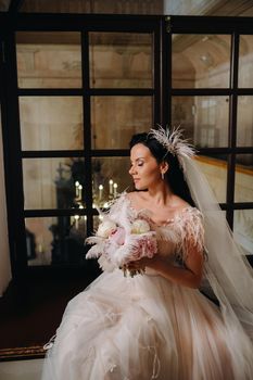 A bride in a wedding dress and a bouquet sits at an open old window and looks.