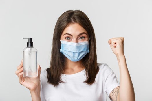 Covid-19, health and social distancing concept. Close-up of attractive smiling girl in medical mask, showing hand sanitizer, recommend antiseptic.