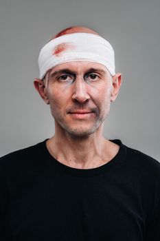 Against a gray background stands a battered and battered man in a black T-shirt with his head wrapped in a bandage.