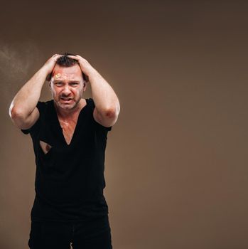 Against a gray background stands a battered angry man in a black T-shirt with wounds.