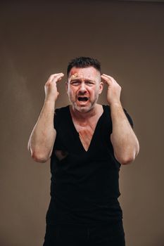 Against a gray background stands a battered angry man in a black T-shirt with wounds.
