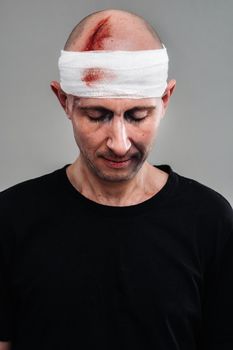 Against a gray background stands a battered and battered man in a black T-shirt with his head wrapped in a bandage.