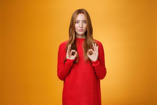 Calm and confident girl showing self-assurance and professionalism as making okay gesture with unemotional expression assuring everything goes by plan standing over orange background, giving approval.