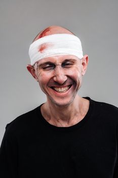 Against a gray background stands a battered and battered man in a black T-shirt with his head wrapped in a bandage.