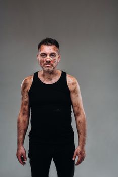 a battered man in a black T shirt who looks like a drug addict and a drunk stands against a gray background.