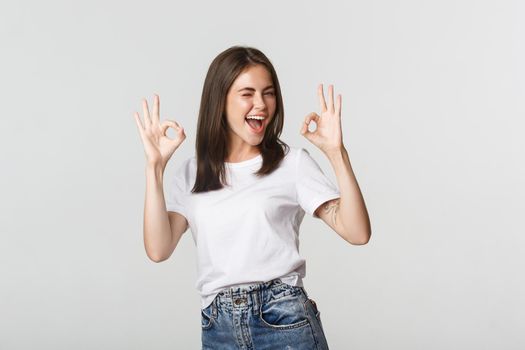 Satisfied cheerful brunette girl winking sassy and showing okay gesture in approval.