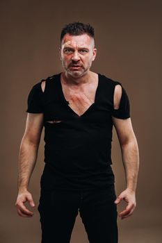 Against a gray background stands a battered angry man in a black T-shirt with wounds.