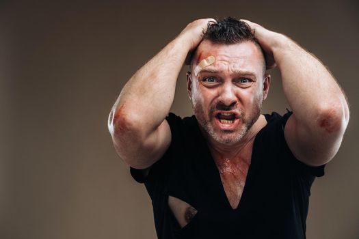 Against a gray background stands a battered angry man in a black T-shirt with wounds.