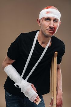 A battered man with a bandaged head and a cast on his arm stands on crutches on a gray background.
