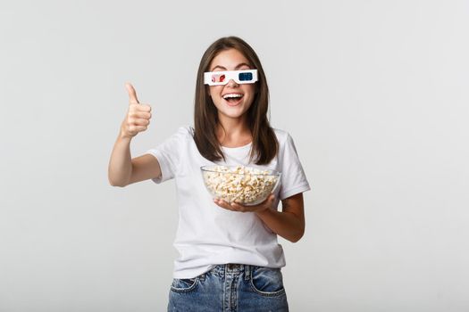 Portrait of satisfied happy girl in 3d glasses, holding bowl with popcorn and showing thumbs-up, like great tv series or movie.