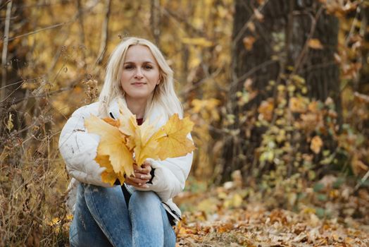 Portrait of a young blonde woman in the autumn forest, with a bouquet of yellow leaves in her hands.