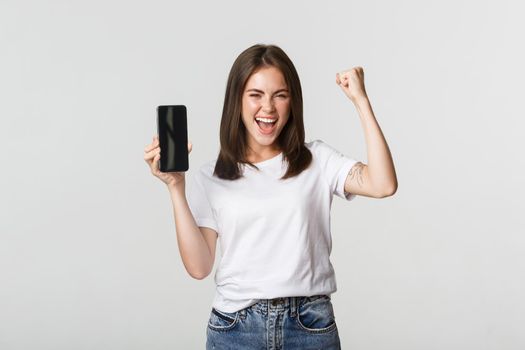 Rejoicing beautiful girl showing smartphone screen and dancing like champion.