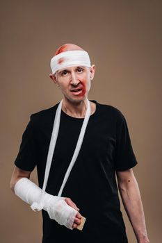 A battered man with a bandaged head and a cast on his arm stands on a gray background.