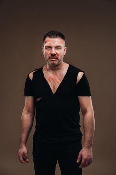 Against a gray background stands a battered angry man in a black T-shirt with wounds.