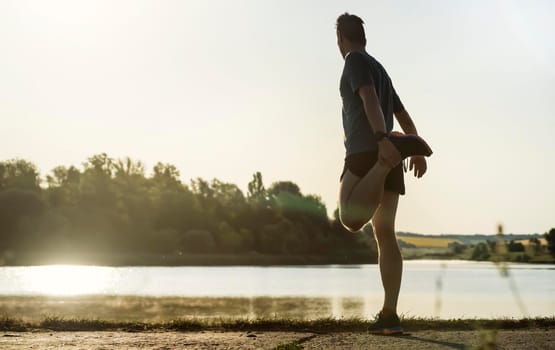 A young man trains outdoors against the backdrop of a beautiful lake at sunset, the athlete warms up, does exercises on the legs before running.