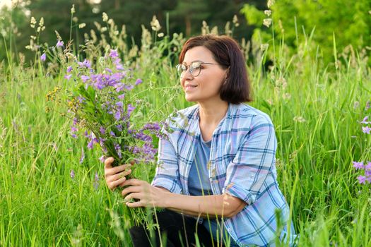 Portrait of middle-aged woman with bouquet of wildflowers in nature. Female 40s age with bells in hands, green spring summer grass meadow background. Beauty, nature, people of mature age concept