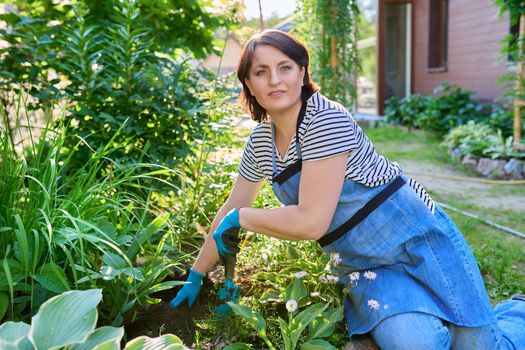 Springtime, spring gardening in backyard. Female in apron of gardening gloves with shovel planting flowering plants in ground from pot. Sunny day green plants, hobbies and leisure of middle aged woman