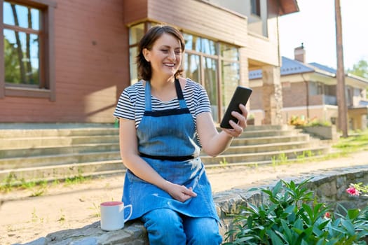 Middle aged woman having video call on smartphone. Happy 40s female sitting in backyard with mug of tea, relaxing on spring summer day, talking online. Lifestyle, video technology, mature people