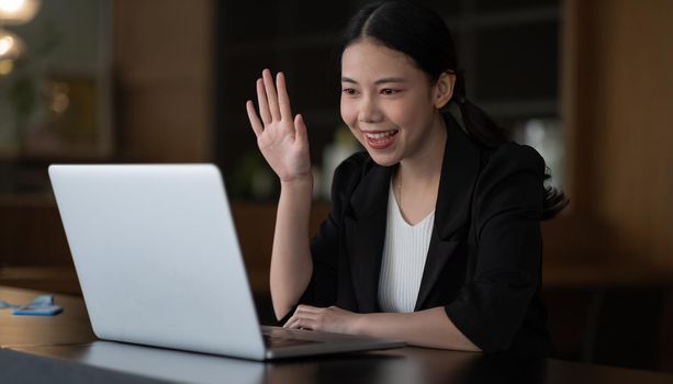 Image of happy asian woman wearing white shirt smiling and waving hand at laptop, while speaking or chatting on video call in office