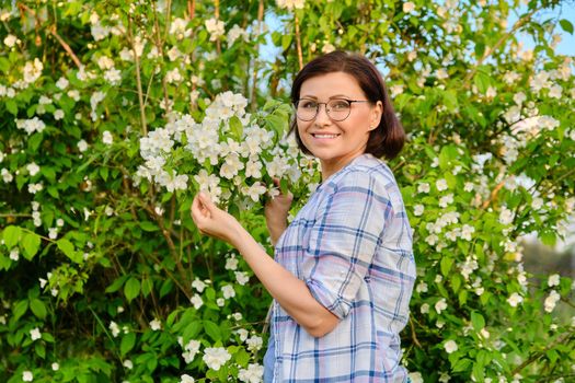 Portrait of smiling middle-aged woman near flowering jasmine bush. Beautiful female 40s of age looking at camera in spring garden. Nature, springtime, beauty, people of mature age concept