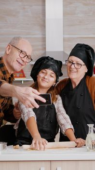 Granddaughter making homemade cookies dough using rolling pin standing at table in xmas decorated culinary kitchen. Happy family smiling while taking selfie enjoying christmas holiday