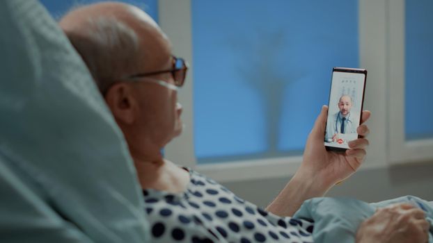 Old patient with disease using smartphone for telemedicine in hospital ward bed at medical unit. Elder man talking with doctor on videocall for illness treatment and professional advice