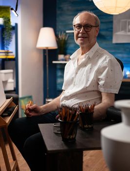 Senior artist looking at camera sitting in workshop at art studio. Caucasian elder person preparing to recreate masterpiece with vase design on table. Old man with drawing creativity