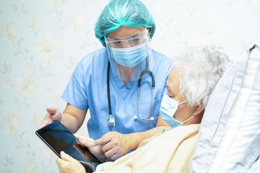 Asian doctor wearing face shield and PPE suit new normal holding tablet with senior woman patient protect safety infection Covid-19 Coronavirus outbreak at quarantine nursing hospital ward.