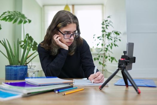 Virtual online lesson, serious teenager guy learns remotely using smartphone on tripod. Video test, meeting, e-education, technology, teaching, adolescence, distance learning, modern college school