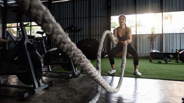 Attractive strong asian fitness woman doing battle ropes exercise at gym. weight training exercise session.