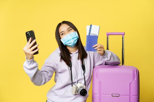 Happy korean woman taking selfie with passport and flight tickets, going on vacation, standing near big cute pink suitcase, yellow background.