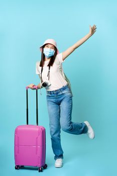 Happy asian girl in medical mask, tourist posing excited with suitcase, going on vacation, travelling abroad, standing over blue background.