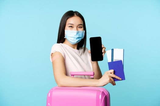 Tourism and healthcare concept. Asian female tourist with suitcare, wearing medical face mask for flight, showing smartphone screen and passport with tickets, vaccination certificate.