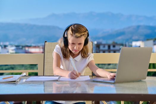 Girl child studying at home online. Pre-teen student in headphones with laptop notebooks sitting on outdoor home terrace. E-education, video chat, virtual lessons, distance learning, technology school